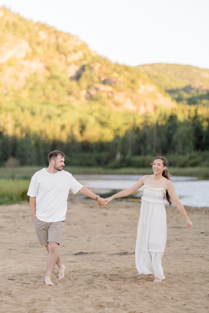 Couple runs hand in hand across a sandy beach with mountain views and autumn foliage in the distance, capturing the destination-inspired atmosphere of Acadia-style engagement photos.