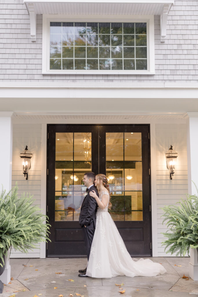 Bride and groom embrace in front of a grand entrance framed by lanterns and symmetrical landscaping after the ceremony, a timeless portrait setting at an elegant Massachusetts reception venue.