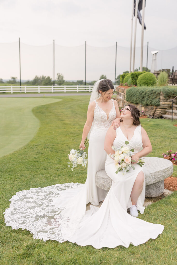 Two brides pose together on a manicured golf course beside layered greenery and soft evening light, reflecting the open scenery often found at unique wedding venues in Massachusetts.