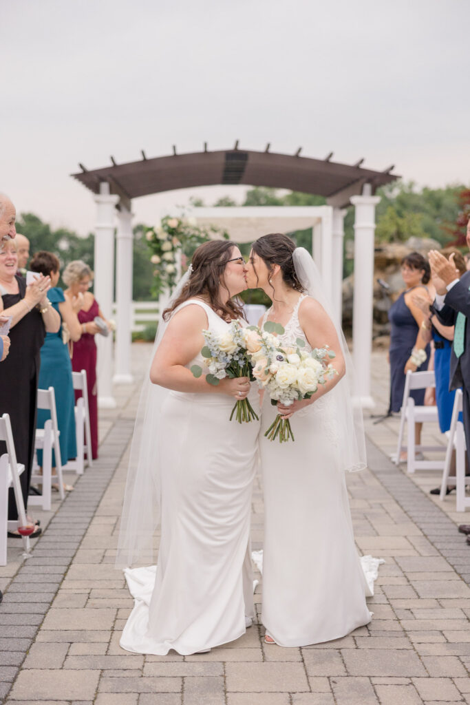 Newly married couple walks back down the aisle together holding bouquets while guests celebrate around them, photographed at a romantic garden wedding venue in Massachusetts.