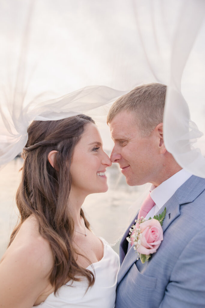 Bride and groom stand forehead to forehead beneath flowing white fabric with the marina softly blurred behind them, capturing the intimate atmosphere of small waterfront wedding venues in Massachusetts.