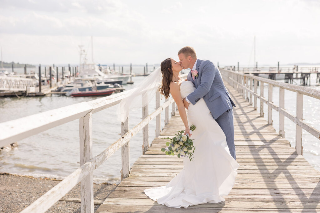 Newly married couple kisses on a weathered dock overlooking sailboats and calm harbor water during sunset portraits, a timeless scene associated with the best waterfront wedding venues Massachusetts couples search for.