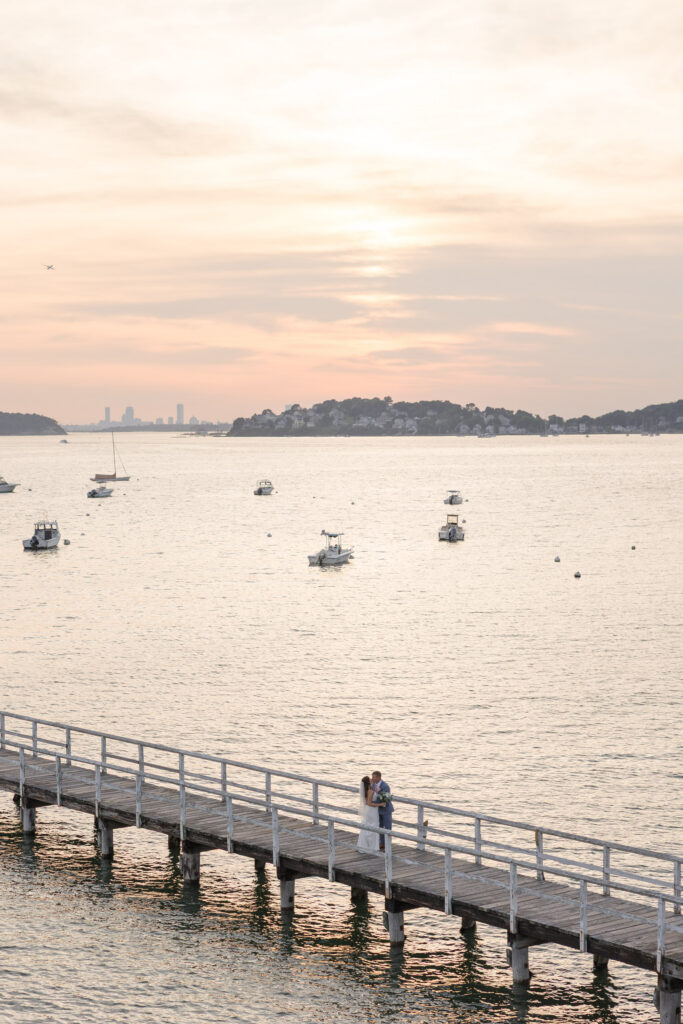 Wide sunset view overlooking calm harbor water and a long wooden pier lined with boats, capturing the peaceful atmosphere that makes many Massachusetts coastal wedding venues feel unforgettable.