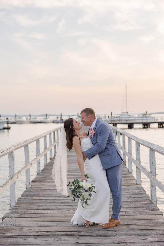 Bride and groom embrace on a weathered dock at sunset with sailboats and harbor views behind them, photographed at one of the best waterfront wedding venues Massachusetts has for coastal celebrations.