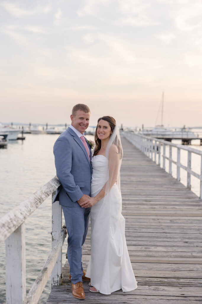 Bride and groom stand together on a weathered dock at sunset overlooking the marina and harbor, a classic scene often associated with coastal Massachusetts wedding venues.