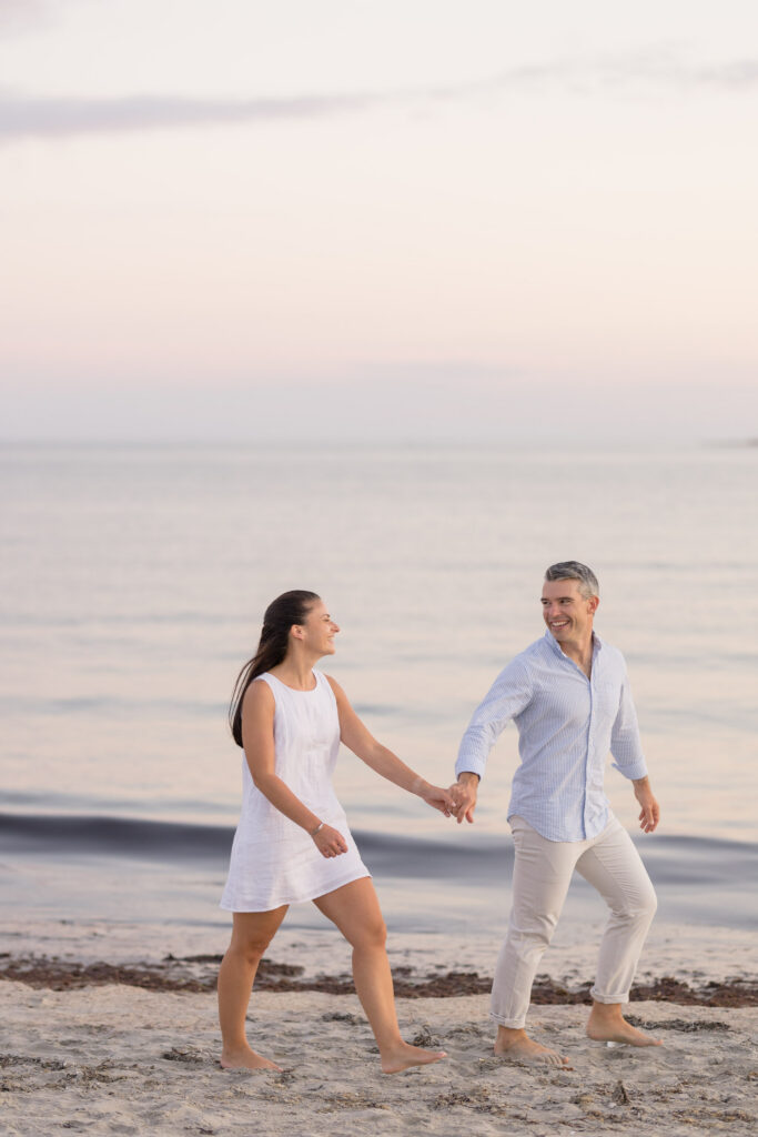 Couple walks barefoot along the shoreline holding hands beneath soft sunset tones and calm ocean water, showcasing the light and candid approach often seen in engagement photographers Massachusetts portfolios.