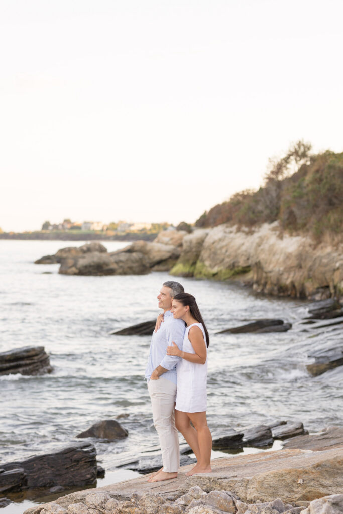 Couple stands together on rocky coastal cliffs overlooking calm water during sunset portraits, reflecting the timeless scenery found in Crane Beach engagement photos and other New England coastal sessions.