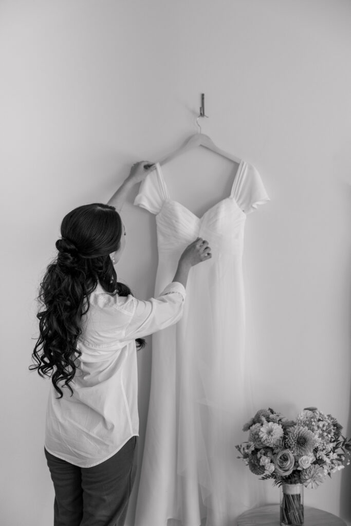 Bridesmaid hangs a minimalist wedding dress against a bright wall beside a bouquet before the ceremony, documenting one of the quiet preparation moments often included in Massachusetts wedding photographer coverage.