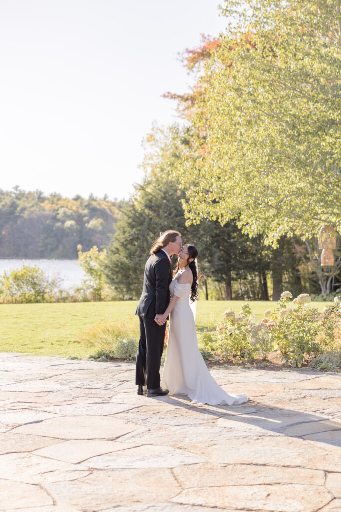 Couple embraces beside a lakeside stone patio surrounded by trees in early autumn light, showcasing the quieter scenery of lakeside wedding venues in Massachusetts.