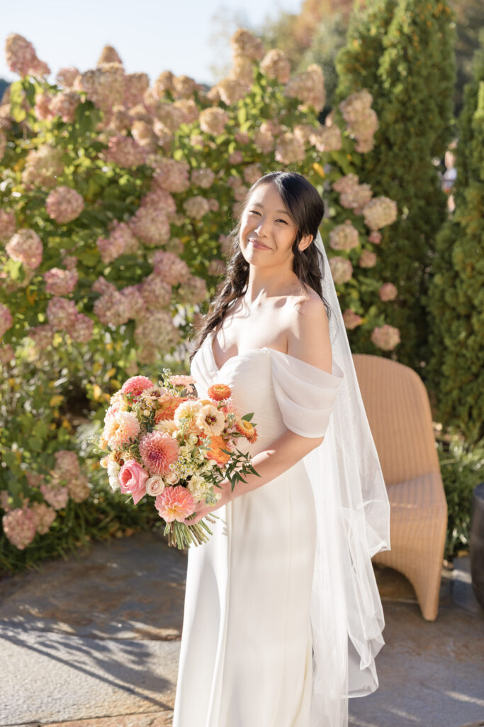 Bride holds a colorful bouquet while standing in front of blooming hydrangeas during golden hour portraits at a garden-inspired waterfront wedding venue in Massachusetts.