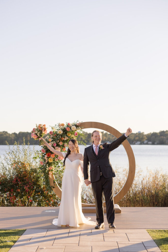 Bride and groom celebrate together beside a lakeside ceremony backdrop framed with florals and a sculptural arch, highlighting romantic outdoor wedding venues Massachusetts couples often love.
