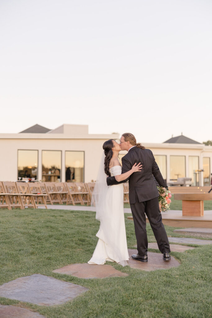 Newly married couple shares a kiss outdoors beside a contemporary reception space with open lawn seating and coastal views, reflecting the atmosphere of luxury waterfront wedding venues Massachusetts couples often search for.