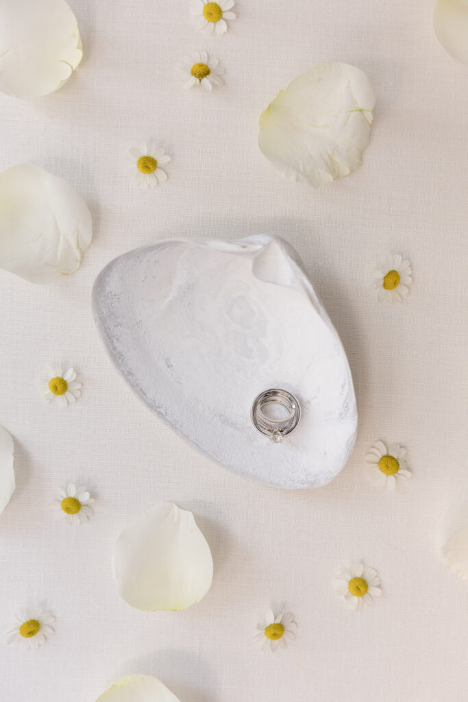 Flat lay of wedding rings styled on a shell dish surrounded by scattered petals and tiny flowers, inspired by the coastal elegance often woven into Cape Cod wedding photography.