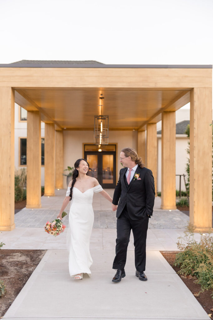 Bride and groom walk hand in hand beneath a modern covered entrance at sunset after their reception, showcasing the clean architecture found at some of the more unique wedding reception venues in Massachusetts.