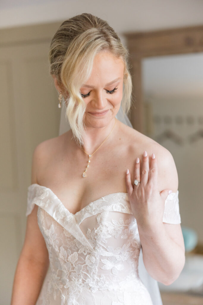 Bride smiles softly while adjusting her necklace in natural window light before the ceremony, photographed with the clean and elegant aesthetic associated with luxury wedding photographers in Massachusetts.
