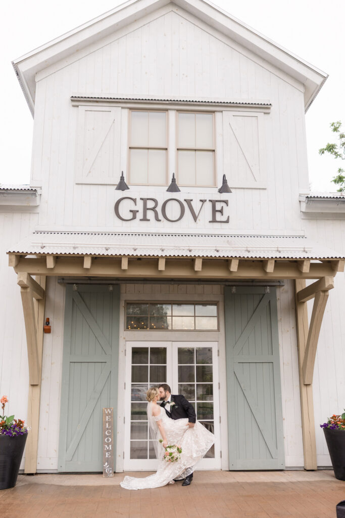 Bride and groom kiss outside a white barn-style venue entrance beneath the Grove sign after the ceremony, showcasing the timeless appeal of barn wedding venues in Massachusetts.