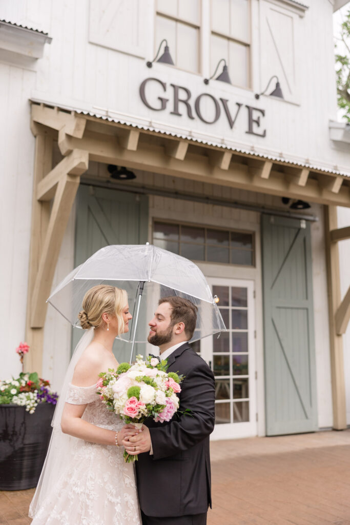 Bride and groom stand beneath a clear umbrella outside a white barn-style venue entrance with soft pastel florals, showcasing the charm of barn wedding venues in Massachusetts with a refined aesthetic.