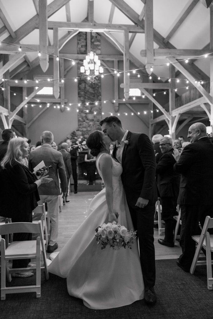 Newly married couple shares a kiss during their ceremony recessional inside a timber-frame venue lit with chandeliers and string lights, capturing the documentary storytelling approach used by experienced wedding photographers in Massachusetts.