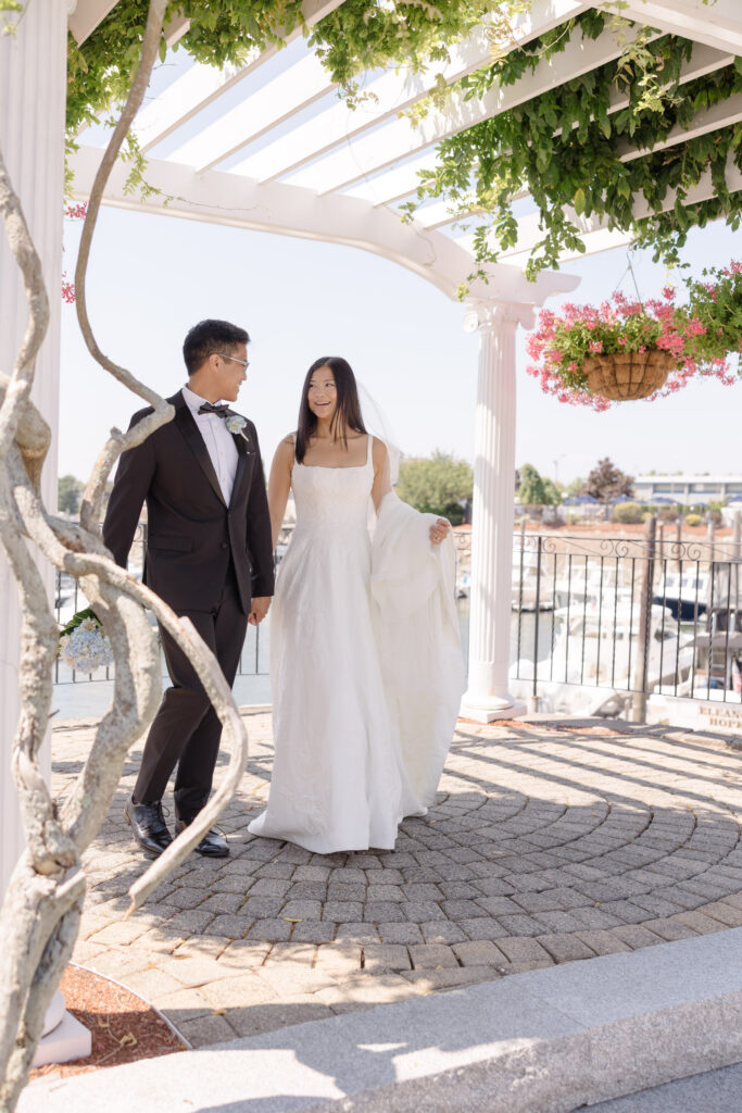 Bride and groom walk beneath a white pergola overlooking the marina during golden hour portraits, photographed at a romantic coastal hotel wedding venue in Massachusetts.