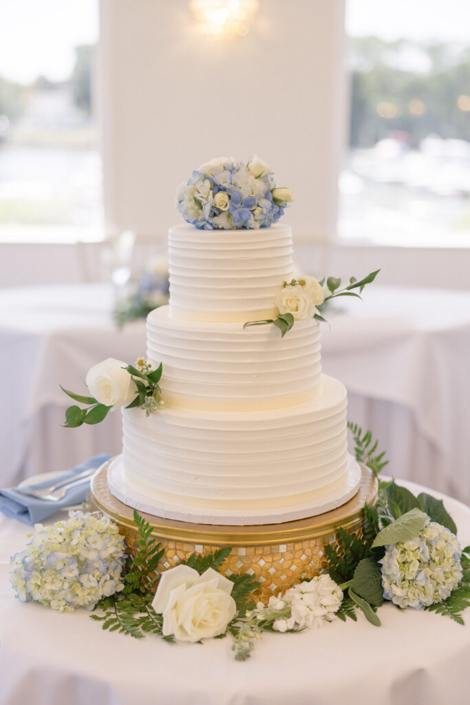 Three-tier white wedding cake decorated with blue hydrangeas and fresh greenery displayed inside a softly lit reception space at an elegant Massachusetts wedding venue.