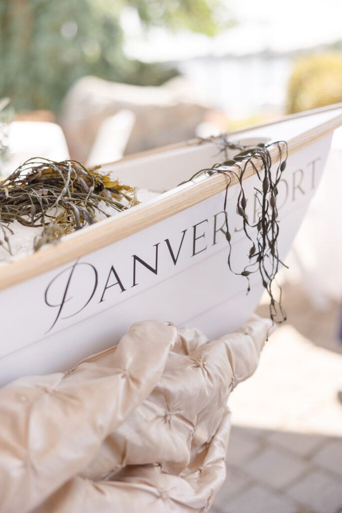 Nautical-inspired welcome display with oyster shells and soft neutral fabrics styled beside a boat marked “Danversport,” highlighting details often found at Gloucester MA wedding venues and harborfront celebrations.