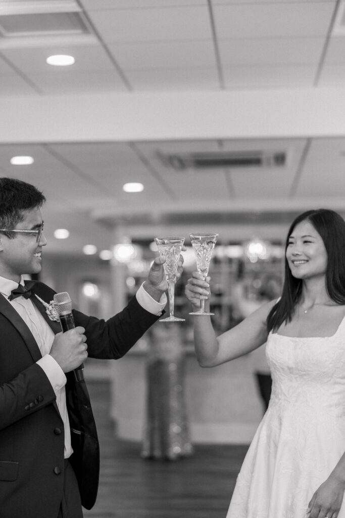 Couple raises champagne glasses together during their reception entrance in a candid black-and-white photograph, highlighting the authentic guest interaction often prioritized in documentary wedding photography Massachusetts coverage.