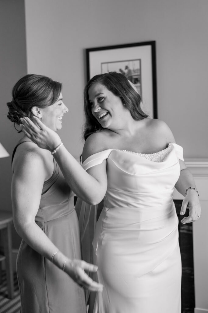 Bride shares a joyful moment with a bridesmaid indoors before the ceremony in a black-and-white candid portrait, reflecting the documentary storytelling style many New England wedding photographers are known for.