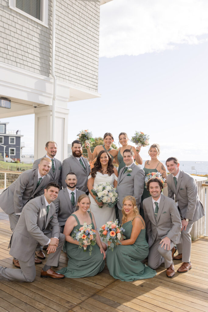 Wedding party gathers on a seaside deck outside a shingled venue with soft neutral tones and ocean air, photographed at one of the memorable wedding venues Massachusetts has along the coast.