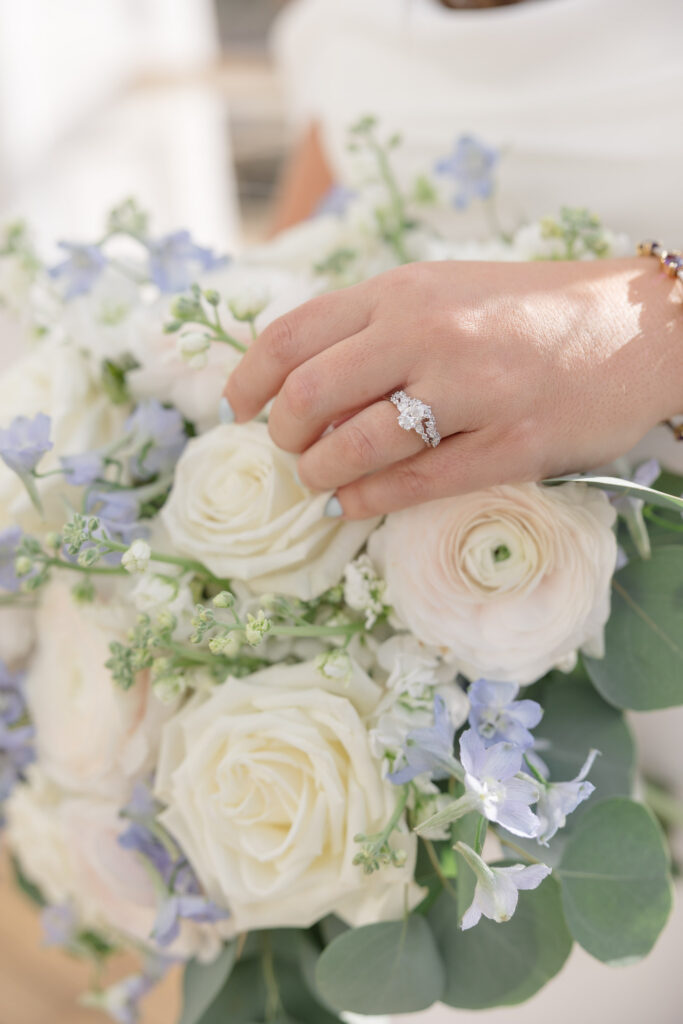Close-up of a bride’s hand resting on a bouquet of white roses and blue florals while showing her engagement ring, photographed with the soft detail work often associated with luxury Cape Cod wedding photographers.