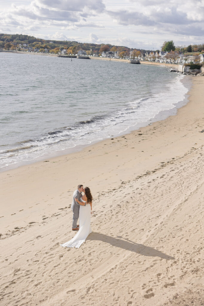 Overhead beach portrait of a bride and groom embracing along the shoreline with soft coastal light and gentle waves behind them, photographed at one of the best waterfront wedding venues Massachusetts has to offer.