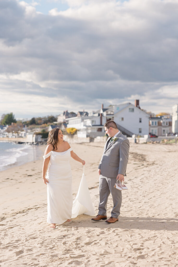 Bride and groom walk barefoot across a sandy beach beside the ocean during sunset portraits, capturing the relaxed atmosphere of coastal Massachusetts wedding venues with guest accommodations nearby.