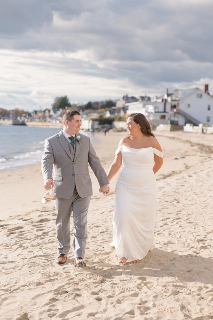 Bride and groom walk barefoot along the shoreline beside coastal homes during sunset portraits with soft ocean light and drifting clouds, photographed in the timeless style many Cape Cod wedding photographers are known for.