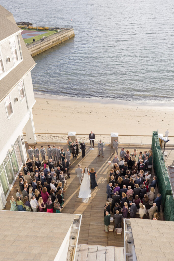 Overhead view of a waterfront ceremony setup with guests gathered beside the beach and ocean beyond, highlighting one of the more unusual Massachusetts wedding venues for a coastal celebration.