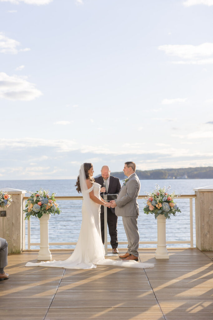 Couple exchanges vows on a wooden ceremony deck overlooking the water and framed with pastel florals, capturing the atmosphere of elegant oceanfront wedding venues in Massachusetts.