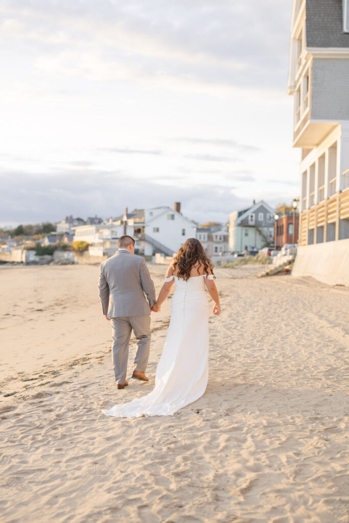 Bride and groom walk barefoot along the beach beside coastal homes during sunset portraits after the ceremony, reflecting the relaxed charm of North Shore Massachusetts wedding venues.