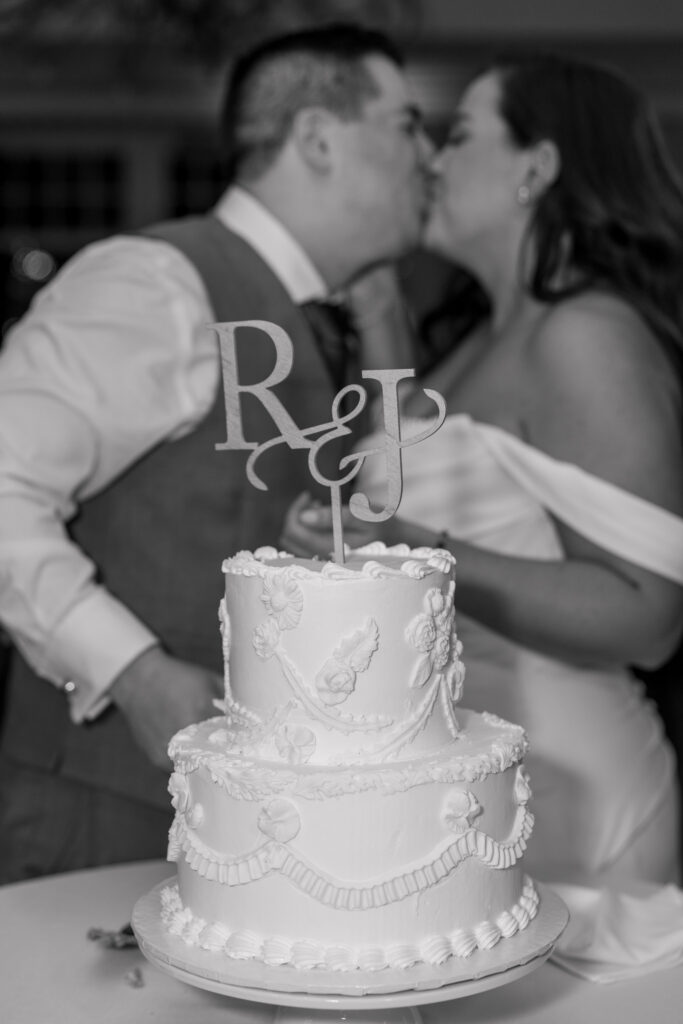 Black-and-white reception portrait of a couple kissing behind a tiered wedding cake with a monogram topper during the reception, photographed at one of the elegant full-service wedding venues in Massachusetts.