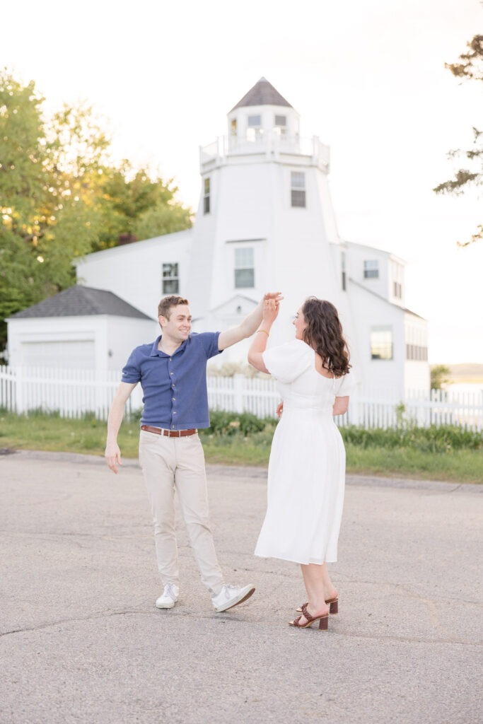 Couple twirls together in front of a white coastal lighthouse during sunset portraits on the North Shore, capturing the charm of Massachusetts engagement photo locations with classic New England character.