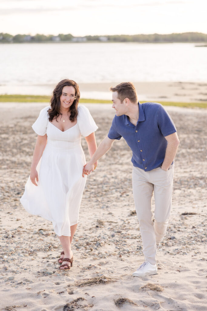 Couple walks hand in hand across a sandy shoreline during golden hour in coordinated neutral outfits, capturing the natural feel often associated with Massachusetts engagement photos.