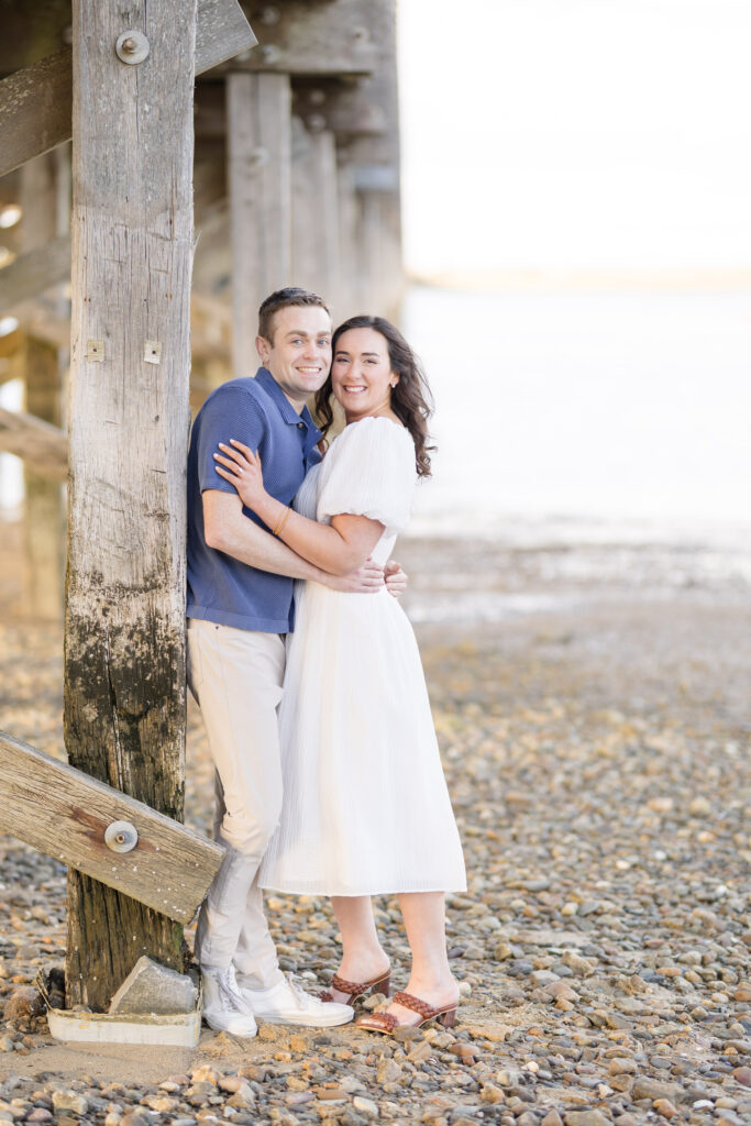 Couple embraces beneath a weathered wooden pier on a rocky beach during golden hour portraits, showcasing one of the more intimate engagement photo locations in Massachusetts.