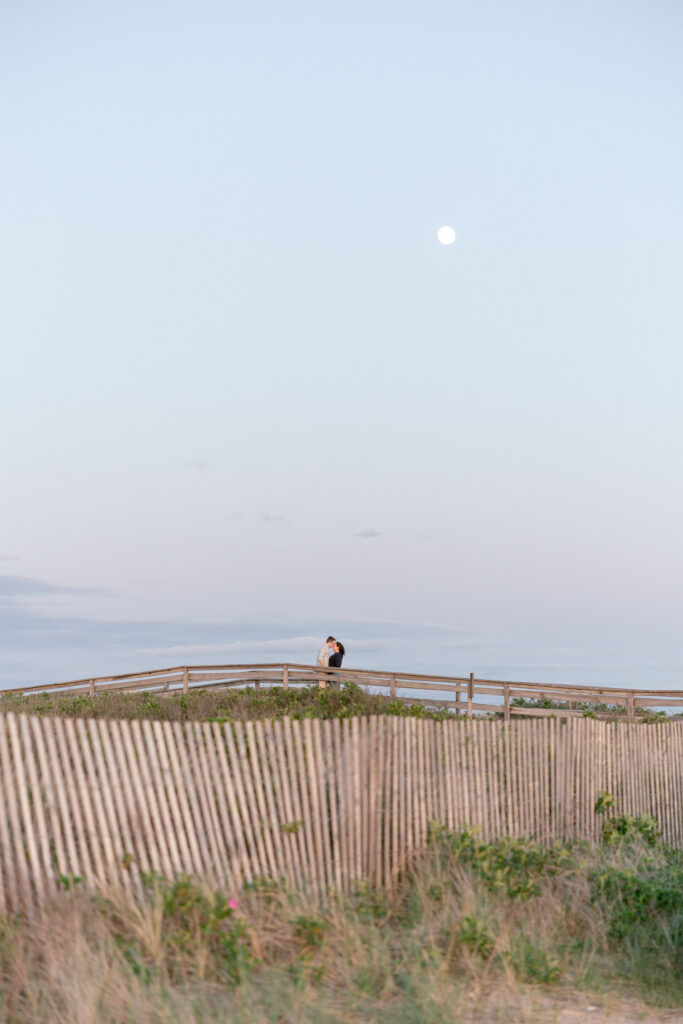 Wide landscape image of a beach boardwalk and dunes beneath a pale evening sky with a couple visible in the distance, showcasing the open scenery of Duxbury Beach engagement photos.