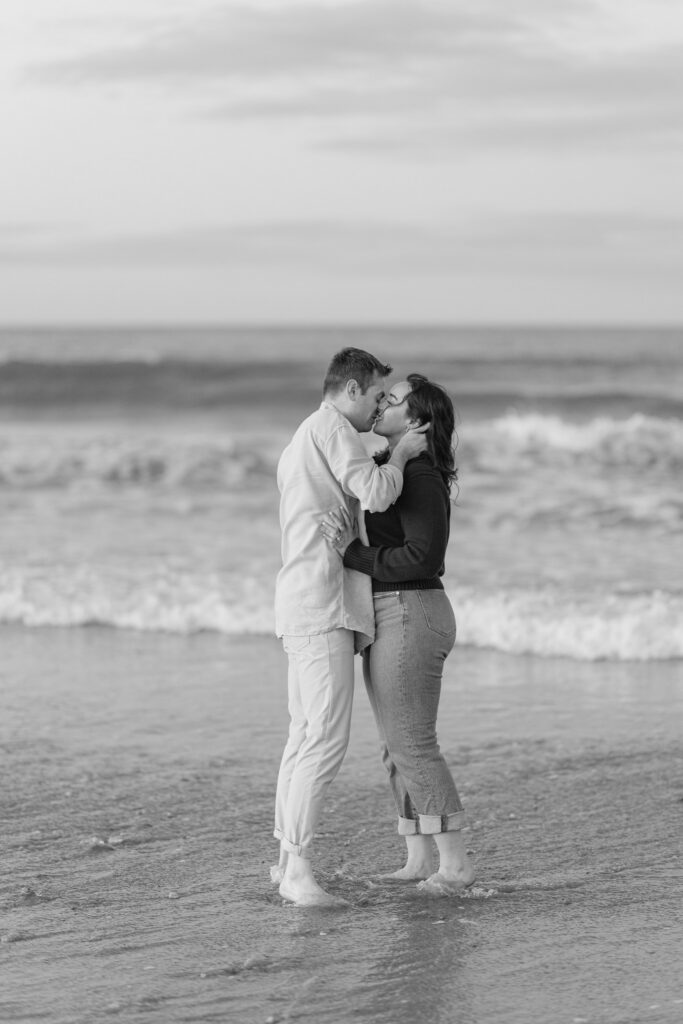 Couple embraces barefoot in the ocean during a black-and-white beach portrait with soft waves rolling in behind them, photographed in the relaxed style many engagement photographers Massachusetts couples love.