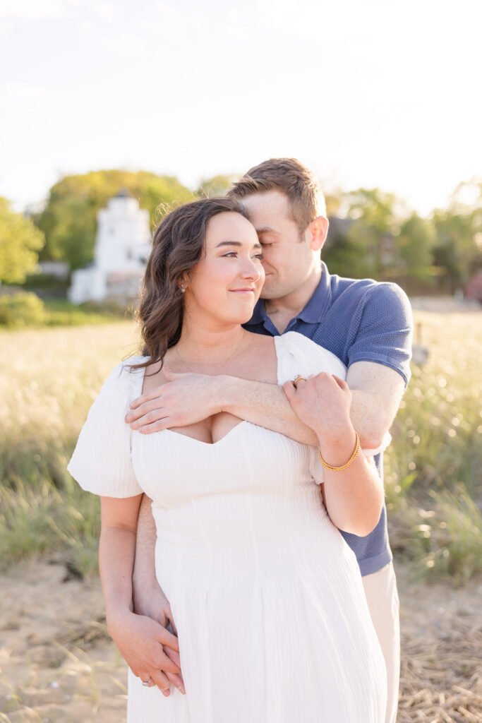 Couple embraces in tall beach grass with a lighthouse softly blurred in the background during golden hour portraits, photographed at one of the more iconic engagement photo locations Massachusetts couples love.