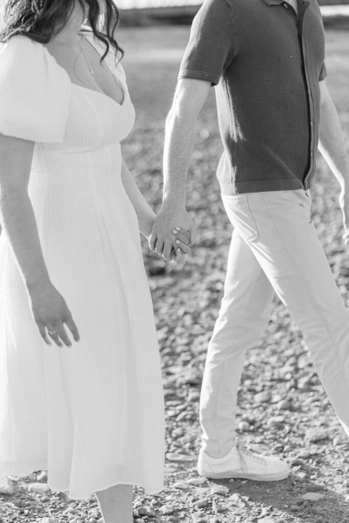 Black-and-white close-up of a couple holding hands while walking along a rocky beach path during an intimate engagement session, highlighting the candid detail work often included in engagement photos Massachusetts galleries.