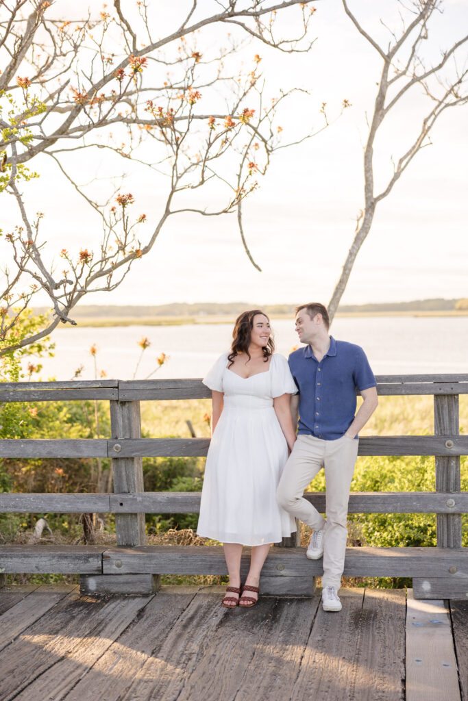 Couple stands together on a wooden boardwalk overlooking the water during golden hour with soft coastal light filtering through the trees, photographed at one of the more scenic engagement photo locations Massachusetts offers.