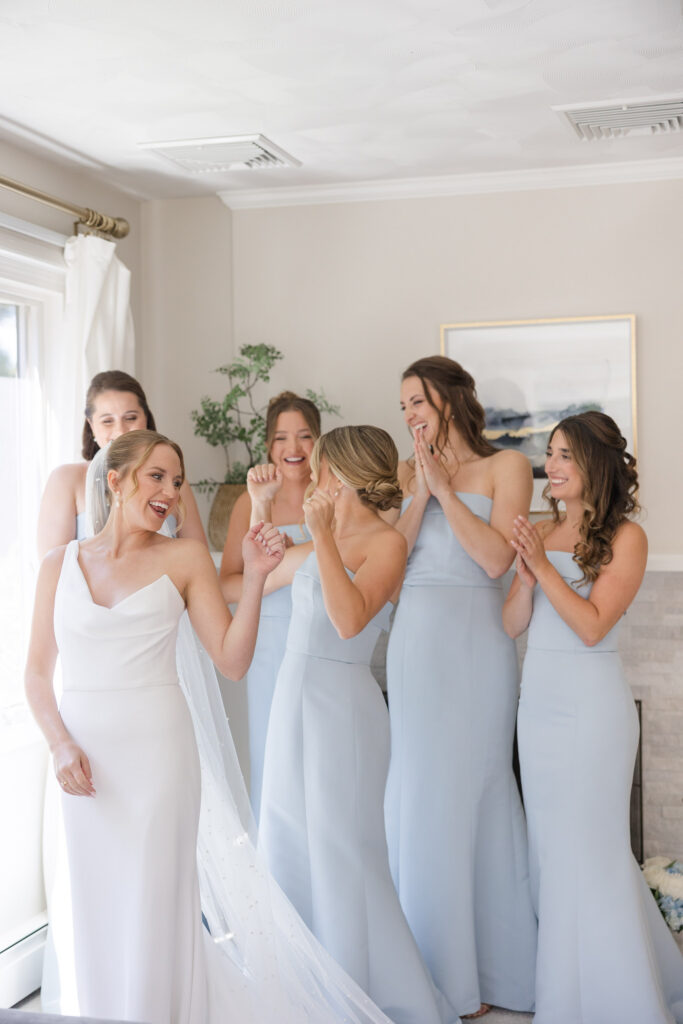 Bride and bridesmaids laugh together during wedding preparations in a softly lit bedroom, highlighting the candid and relaxed moments that make wedding photography in Massachusetts feel personal and true to life.