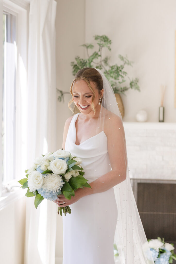 Bride stands in soft natural window light holding a bouquet of white blooms before the ceremony, photographed with the airy and romantic aesthetic many Cape Cod wedding photographers are known for.