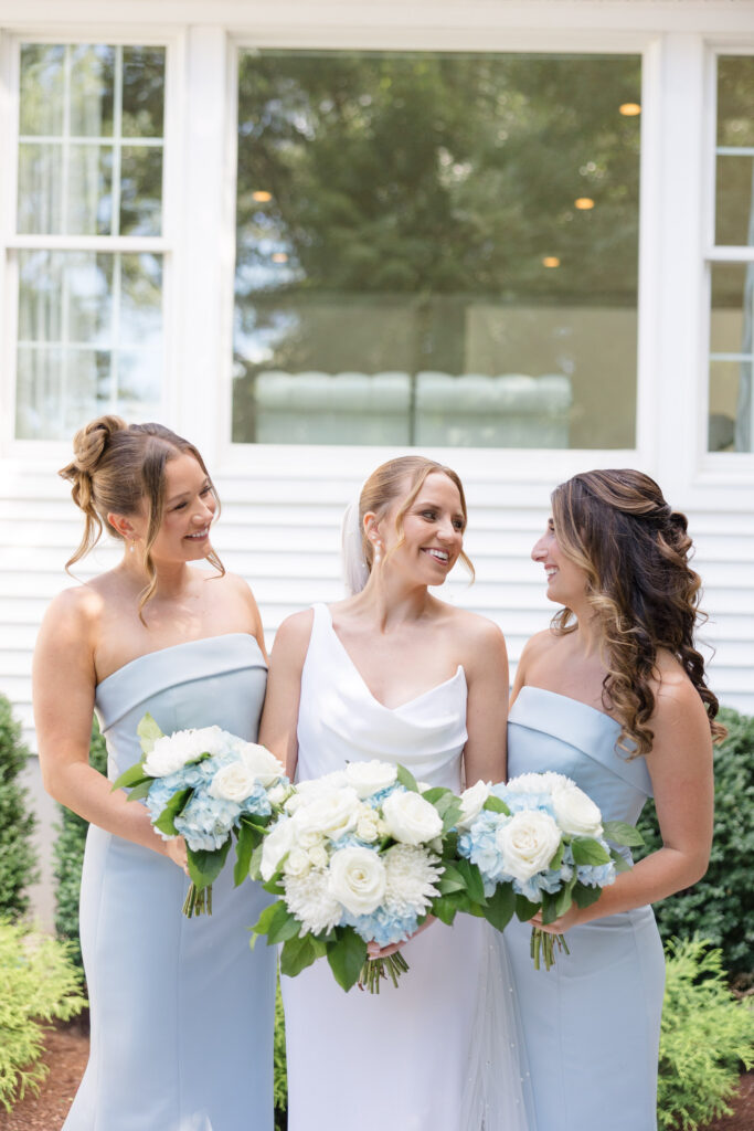 Bride and bridesmaids stand together outside in pale blue dresses holding white floral bouquets during pre-ceremony portraits, a timeless bridal party moment often featured in Massachusetts wedding photography galleries.