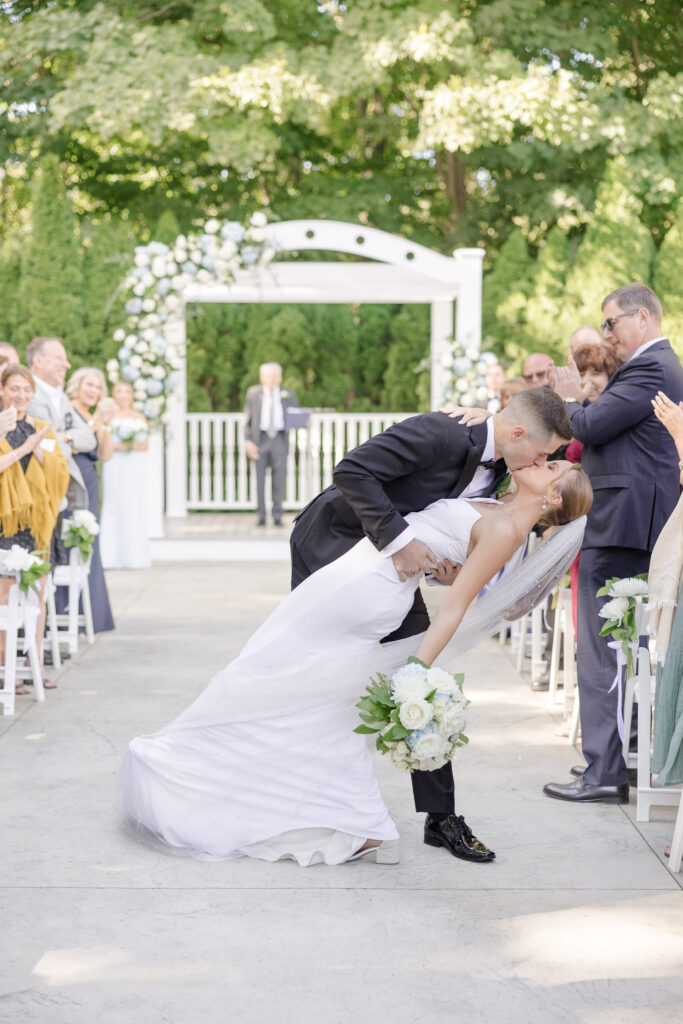 Groom dips the bride during the ceremony recessional while guests cheer beneath a floral arbor, capturing the joyful energy often associated with outdoor wedding venues in Massachusetts.