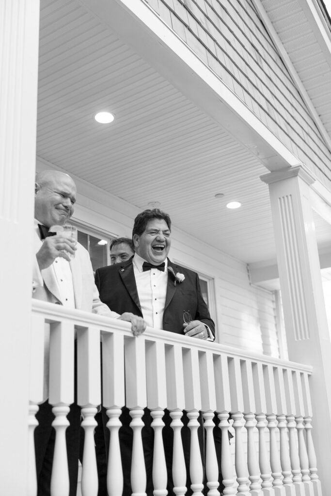 Groom laughs with family members from an upper porch during cocktail hour in a candid black-and-white image, capturing the relaxed guest experience found at many Massachusetts wedding venues.