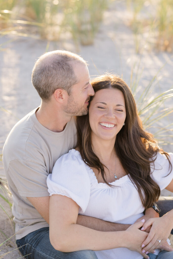 Man kisses his partner on the cheek while they sit together in the dunes surrounded by beach grass and warm evening light, photographed in the soft editorial style many Massachusetts engagement photographers are known for.